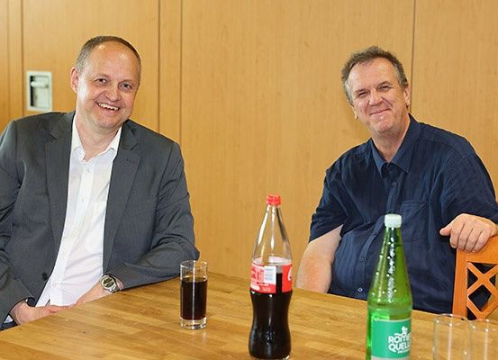 Zwei Männer sitzen an einem Tisch mit Getränken, einer in einem Anzug und der andere in einem blauen Hemd. Hinter ihnen ist eine Holzwand.