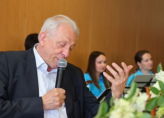 Ein älterer Mann spricht in ein Mikrofon bei einer formellen Veranstaltung, mit zwei Frauen in Blau hinter ihm.