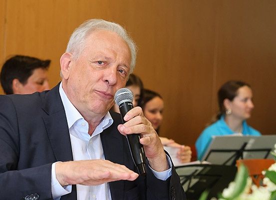 Ein älterer Mann in einem Anzug spricht bei einer Veranstaltung in ein Mikrofon. Hinter ihm sitzen Leute, eine Frau trägt ein blaues Hemd.