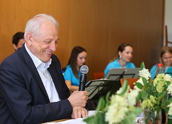 Ein älterer Mann sitzt mit einem Mikrofon am Tisch, lächelt und ist von Blumen und sitzenden Frauen umgeben.