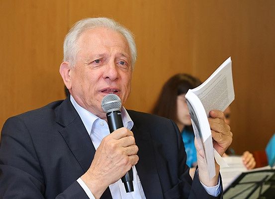 Ein älterer Mann in einem Anzug spricht in ein Mikrofon, während er ein Buch in der Hand hält. Hinter ihm schaut eine Frau auf ein Papier. Eine Holzwand und eine weitere Person mit einem blauen Shirt sind im Hintergrund zu sehen.