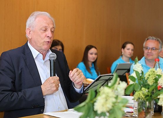 Ein älterer Mann in einem Anzug spricht in ein Mikrofon bei einer Versammlung mit anderen, die hinter ihm sitzen.