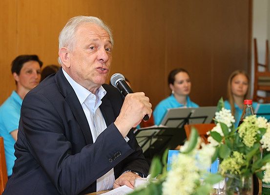 Ein älterer Mann in einem Anzug spricht in ein Mikrofon bei einer Besprechung. Auf dem Tisch vor ihm stehen Blumen und Flaschen. Hinter ihm sitzen mehrere Personen und schauen ihn an.