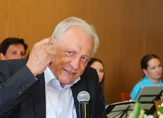 Ein älterer Mann in einem dunklen Anzug lächelt und gestikuliert mit seiner Hand, während er in ein Mikrofon spricht bei einer Veranstaltung. Dahinter ein verschwommenes Bild einer Frau mit einer Musikpartitur und Blumen.