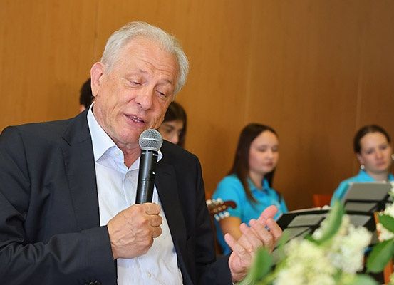 Ein älterer Mann in einem Anzug spricht in ein Mikrofon. Hinter ihm sitzen drei Frauen, eine spielt Gitarre und eine andere liest Noten. Der Raum hat Holzwände und eine Blumendekoration im Vordergrund.