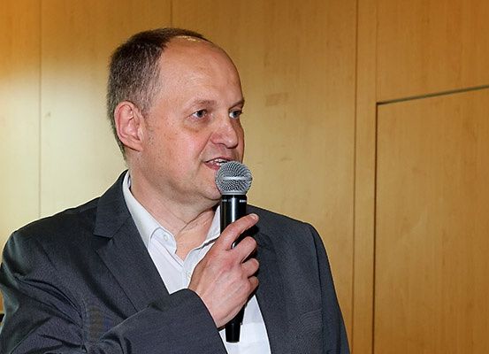 Ein Mann in einem dunklen Anzug spricht in ein Mikrofon, möglicherweise auf einer Konferenz. Die Wand hinter ihm ist aus Holz.