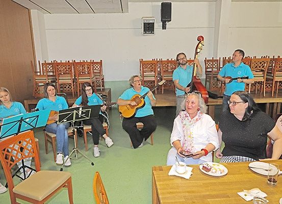 Eine Gruppe Erwachsener in blauen Shirts spielt Gitarren in einem Raum mit einem Holztisch und Stühlen.