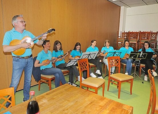 Ein Mann spielt Gitarre, während eine Gruppe von Frauen um ihn herumsitzt, Gitarren spielt und Noten liest. Sie sind in einem Raum mit Holzwänden und einem Holztisch.