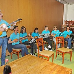 Ein Mann spielt Gitarre, während eine Gruppe von Frauen um ihn herumsitzt, Gitarren spielt und Noten liest. Sie sind in einem Raum mit Holzwänden und einem Holztisch.