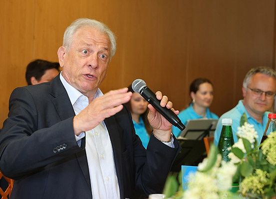 Ein älterer Mann spricht in ein Mikrofon und gestikuliert mit der Hand. Hinter ihm sitzen eine Frau und ein Mann. Auf dem Tisch steht eine Blumenvase.