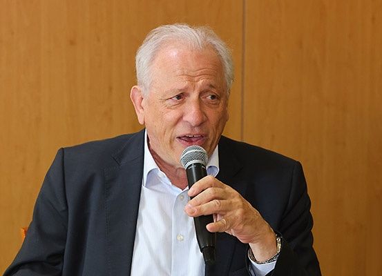 Ein älterer Mann sitzt auf einem Stuhl, hält ein Mikrofon in der Hand und scheint eine Rede zu halten. Er trägt einen dunklen Anzug, ein weißes Hemd und hat weißes Haar.