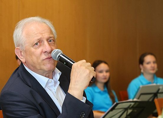 Ein älterer Mann in einem Anzug hält ein Mikrofon und spricht zu einer Gruppe. Zwei Frauen sitzen hinter ihm, eine mit Notenblättern.