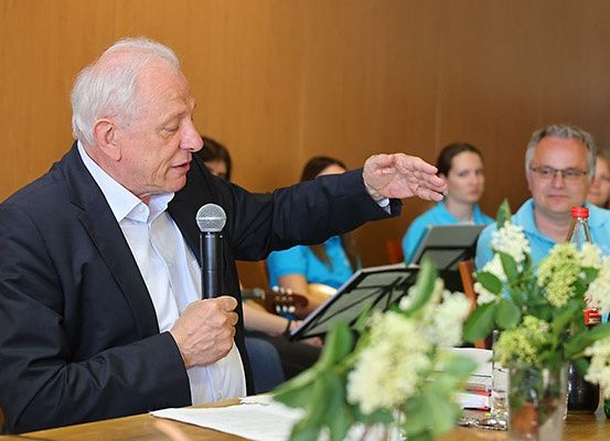 Ein älterer Mann spricht in ein Mikrofon bei einer Konferenz. Hinter ihm spielt eine Frau Gitarre. Andere sitzen und lauschen. Ein Tisch mit Blumen und einer Flasche ist im Vordergrund.