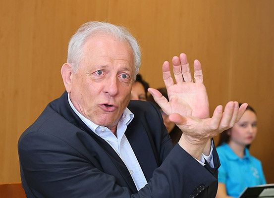 Ein älterer Mann in einem schwarzen Anzug macht Gesten, während er spricht, wahrscheinlich in einer Besprechung. Hinter ihm sitzen zwei Frauen, eine mit einem blauen Shirt.