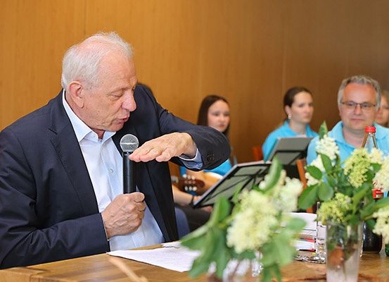 Ein älterer Mann sitzt an einem Tisch, spricht in ein Mikrofon und gestikuliert mit der Hand. Hinter ihm sitzen andere Personen, von denen eine eine Gitarre spielt.
