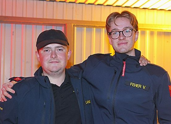 Zwei Männer in dunkelblauen Jacken mit roten Aufnähern und gelbem Text posieren für ein Foto. Einer trägt einen schwarzen Hut, der andere eine Brille.