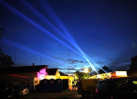 Nachtansicht eines Gebäudes mit blauen und rosafarbenen Lichtern, umgeben von geparkten Autos und Straßenlaternen, mit Lichtstrahlen am Himmel.