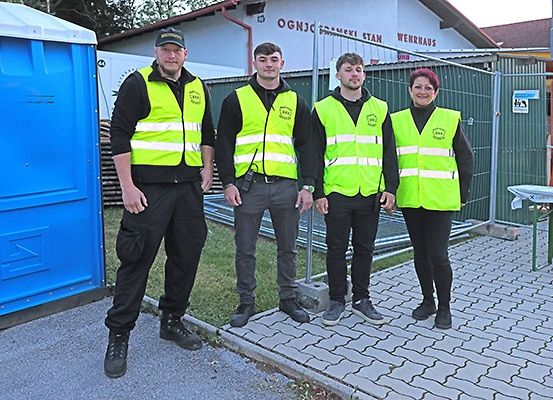 Vier Personen in reflektierenden Westen stehen auf einem Betonweg und posieren für ein Foto vor einem Gebäude mit einem Schild, das 'OGNJC' liest.