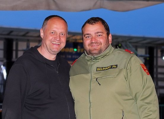 Zwei Männer lächeln für ein Foto bei einer Outdoor-Veranstaltung. Ein Mann trägt eine schwarze Kapuzenjacke, der andere eine grüne Jacke mit 'BELIHART PH' auf der Brust.