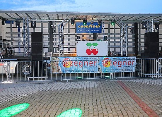 Eine Bühnenaufbau mit Lautsprechern, Beleuchtung und Bannern, einschließlich eines mit einem Kirschenlogo. Der Bereich ist eingezäunt.