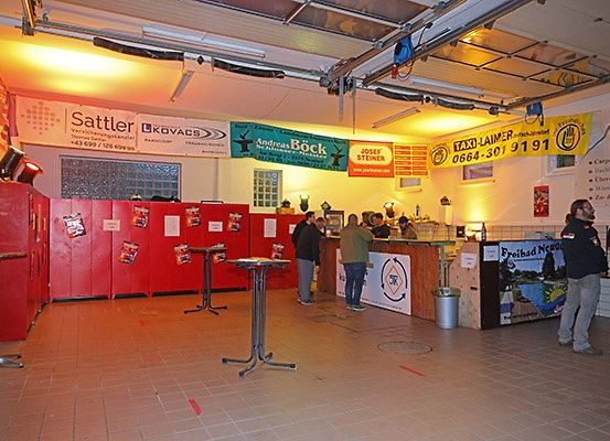 Eine Gruppe von Menschen steht in der Nähe einer Theke in einer Einrichtung. Werbebanner hängen an der Decke. Ein Mülleimer steht in der Nähe der Theke.