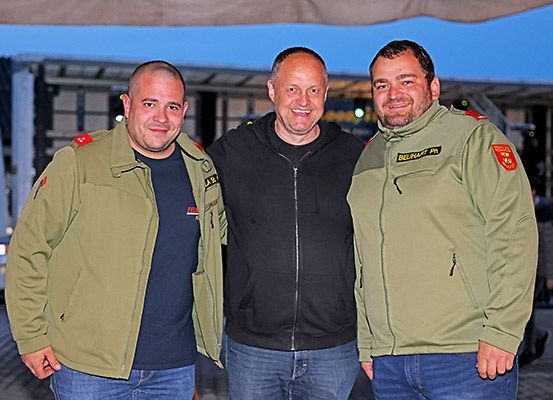 Drei Männer stehen zusammen, einer in einer olivgrünen Militärjacke, ein anderer in einer schwarzen Kapuzenjacke und der dritte in einer grünen Militärjacke, alle lächeln für ein Foto.