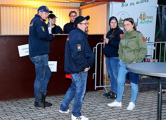 Mehrere Personen in Uniform stehen vor einem Tresen mit Schildern, zwei unterhalten sich, während andere zusehen; ein Tisch und eine Metallbarriere sind ebenfalls sichtbar.