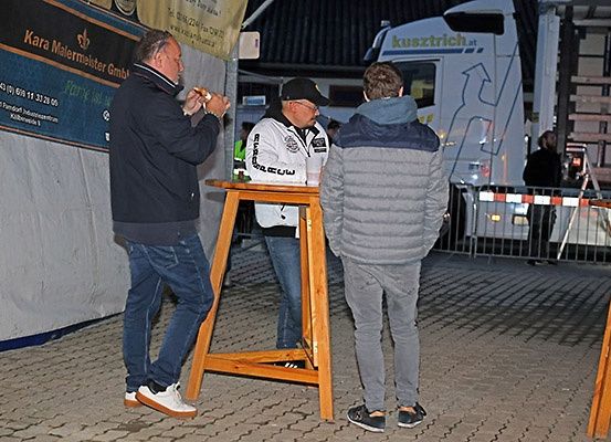 Drei Männer stehen an einer Bar, einer von ihnen raucht eine Zigarette. Dahinter steht ein LKW mit einem Logo an der Seite.