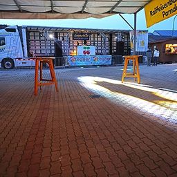 Ein Außenbereich mit einem Ziegelboden, einer Bühne mit einem geparkten weißen LKW, einem Banner und einigen Leuten, die in der Nähe der Bühne und eines kleinen Imbisswagens stehen.