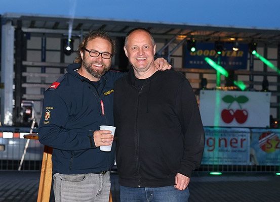 Zwei Männer stehen vor einer Bühne mit grünen Lichtern und einem Banner. Ein Mann hält einen Becher, beide lächeln.