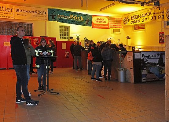 Eine Gruppe von Menschen steht in einer Halle, möglicherweise in einer Warteschlange. Sie tragen Winterkleidung, und einige halten Papiere. Werbebanner hängen an den Wänden, und ein Mülleimer ist in der Nähe platziert.