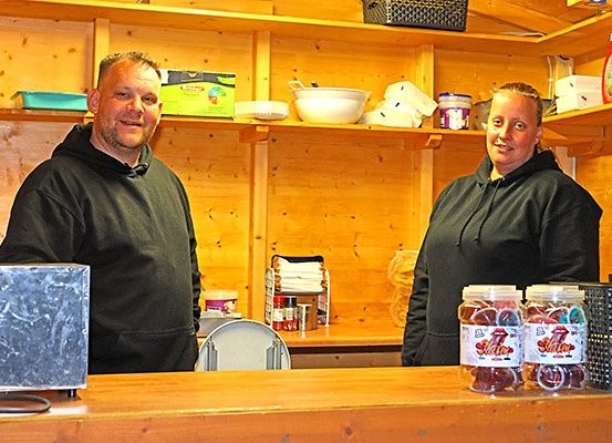 Zwei Personen stehen in einer Küche mit Holzregalen und Gläsern mit Süßigkeiten auf der Theke. Der Mann trägt eine schwarze Kapuzenjacke und die Frau hat ein lächelndes Gesicht. Hinter ihnen befinden sich Küchenutensilien und Lebensmittelbehälter auf den Regalen.