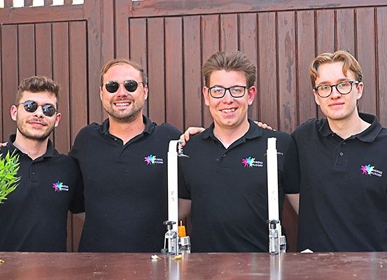 Vier Männer in schwarzen Polo-Shirts stehen hinter einem Tisch, lächeln und posieren für ein Foto. Einer hat eine Topfpflanze auf der linken Seite.