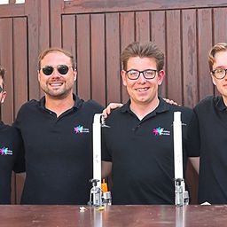 Vier Männer in schwarzen Polo-Shirts stehen hinter einem Tisch, lächeln und posieren für ein Foto. Einer hat eine Topfpflanze auf der linken Seite.