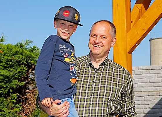 Ein Mann in einem karierten Hemd lächelt, während er einen kleinen Jungen in einem Baseballcap draußen in der Nähe eines Holzbaues und einer Ziegelmauer hält.