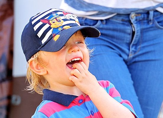 Ein junger Junge trägt eine blau-weiß gestreifte Kappe und ein farbenfrohes Shirt und isst, während jemand in Jeans hinter ihm steht.