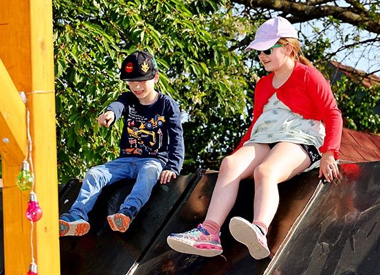 Zwei Kinder, ein Junge und ein Mädchen, sitzen auf einem schwarzen Rutschen in einem Park. Sie tragen Mützen, eine Sonnenbrille und Turnschuhe.