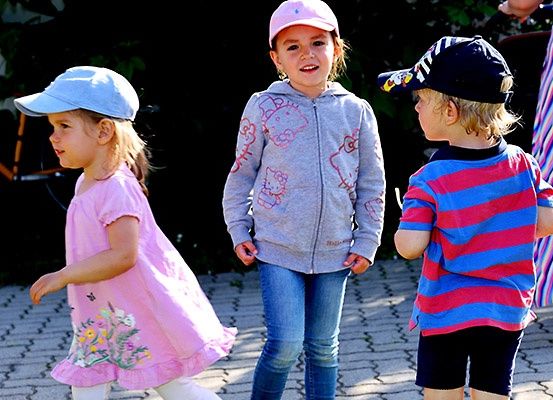 Drei kleine Kinder stehen zusammen auf einem gepflasterten Weg, jedes mit einer Mütze und in unterschiedlich farbiger Kleidung.