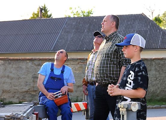 Fünf Männer stehen vor einem Gebäude, einer sitzt auf einem Metallobjekt. Sie alle schauen nach oben, möglicherweise zu einem Flugzeug oder einer Veranstaltung. Ein Mann hält eine Kiste, und ein anderer trägt eine Baseball-Kappe.