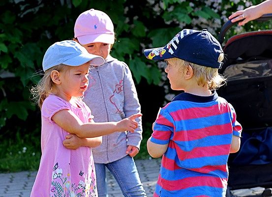 Drei kleine Kinder in Mützen stehen draußen in der Nähe eines Kinderwagens. Ein Mädchen zeigt auf einen Jungen, während ein anderes Mädchen hinter ihr steht.