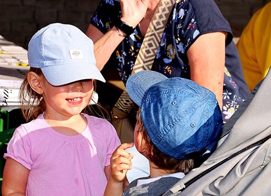Zwei Kinder in Mützen interagieren, eines mit lächelndem Gesicht, in der Nähe eines Kinderwagens. Ein Erwachsener, der ein Blumenhemd trägt, ist im Hintergrund zu sehen.