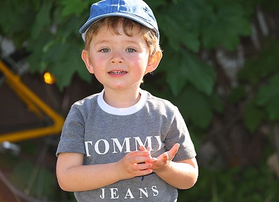 Ein junger Junge in einer blauen Kappe und einem grauen Shirt mit weißem Text 'TOMMY JEANS' steht draußen.