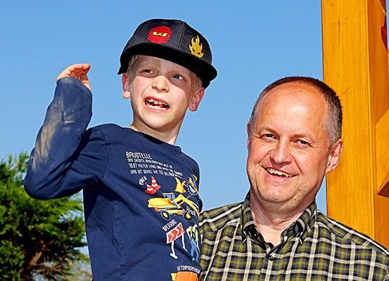 Ein lächelnder Mann und ein junger Junge stehen vor einer Holzstruktur, wobei der Junge ein blaues T-Shirt mit einem Baufahrzeug-Grafik trägt.