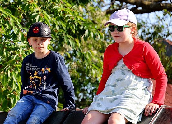 Zwei Kinder, ein Junge und ein Mädchen, sitzen auf einer Metallstruktur, möglicherweise einem Dach. Sie tragen beide eine Sonnenbrille. Der Junge trägt eine Mütze und ein blaues T-Shirt mit einem LKW-Aufdruck. Das Mädchen trägt ein weißes Kleid und eine rote Strickjacke. Hinter ihnen steht ein Baum mit grünen Blättern.
