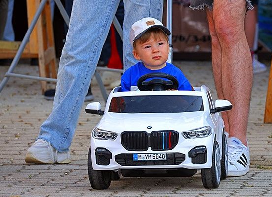 Ein Kleinkind sitzt in einem Spielzeugauto, das einem BMW ähnelt, trägt ein blaues Shirt und eine weiße Kappe. Er schaut nach vorne, mit zwei Personen hinter dem Auto stehend.