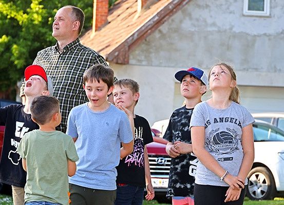 Eine Gruppe von Kindern und ein Mann schauen nach oben auf einem Außenbereich. Sie scheinen etwas zu beobachten. Hinter ihnen befindet sich ein Gebäude mit einer Garage und ein Auto, das davor geparkt ist.