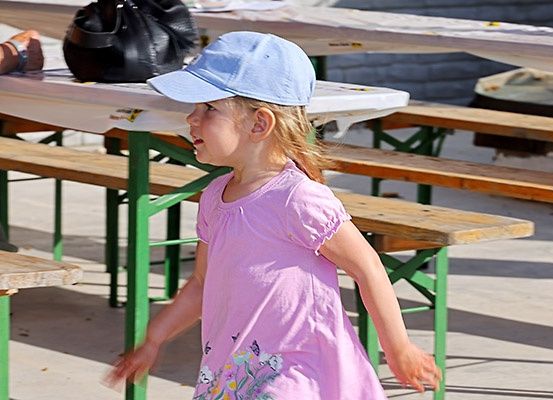 Ein kleines Mädchen in einem rosa Kleid und einer blauen Mütze geht durch einen Park. Hinter ihr sind Holzbänke und ein Tisch mit einer schwarzen Tasche darauf.