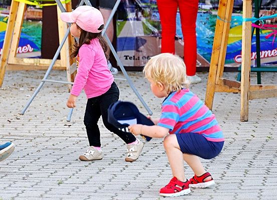 Zwei Kinder spielen mit Hüten auf einem Ziegelweg. Eines im Rosa, das andere in Streifen, mit Metallständern und buntem Hintergrund.