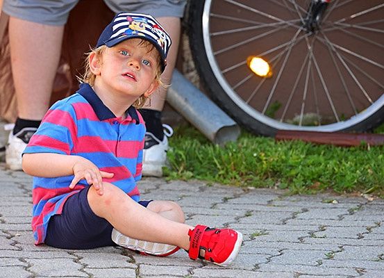 Ein junger Junge mit gestreiftem Shirt und Mütze sitzt auf dem Boden und schaut nach oben. Dahinter sind die Beine von jemandem in Turnschuhen zu sehen. Im Hintergrund befindet sich ein Fahrradreifen mit einer leuchtenden Lampe.
