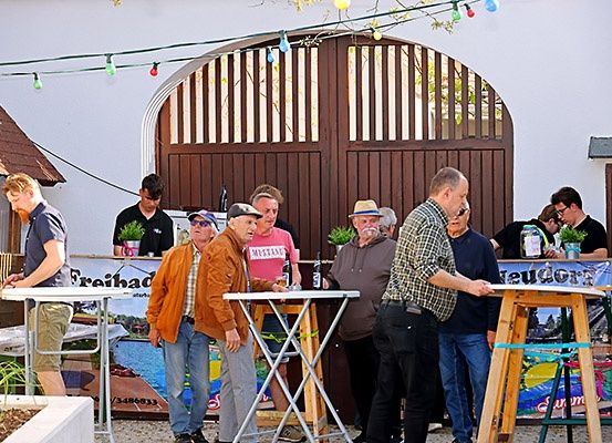 Eine Gruppe von Menschen versammelt sich im Freien in der Nähe einer Bar mit Tischen und Topfpflanzen. Ein Mann steht hinter der Theke.